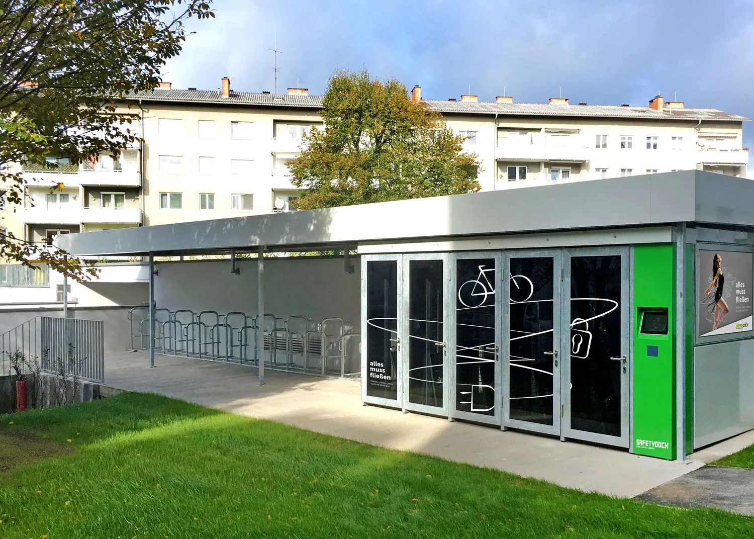 Moderne Mobilitätsstation in Linz-Urfahr mit überdachtem Fahrradabstellplatz und safetydock E-Bike-Ladesystem, umgeben von Wohnhäusern und Grünfläche